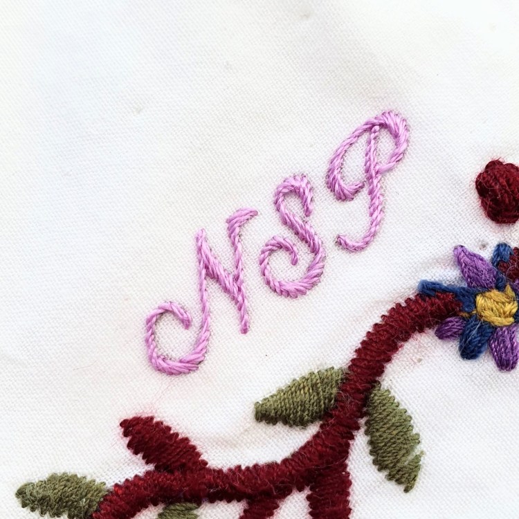 Lilac hand embroidered letters NSP on white fabric with a scrolling border of leaves below it 