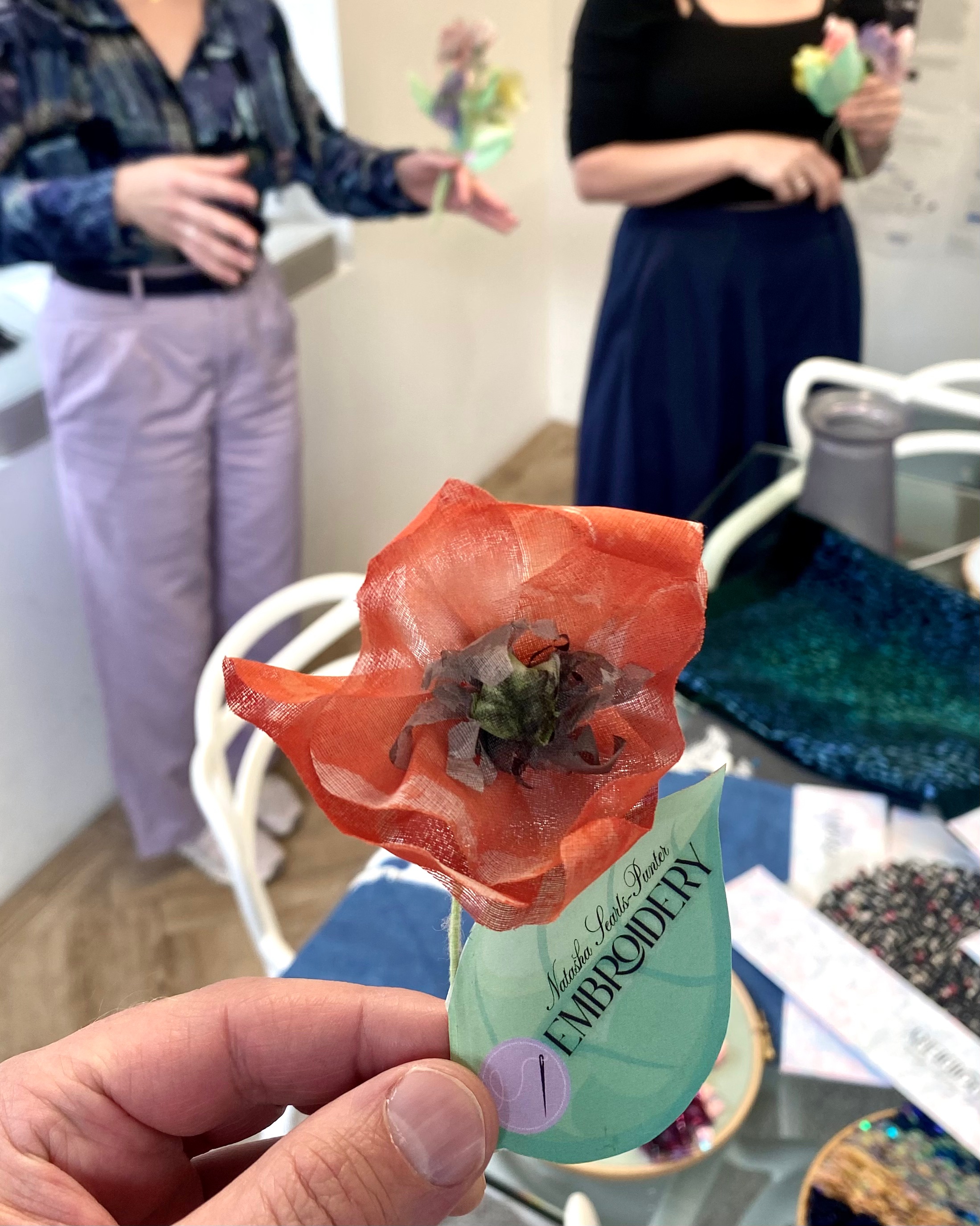 Red silk fabric flower held in a mans hand with Natasha Searls-Punter Embroidery details on the leaf attached to its stem. Women stand out of focus in the background in conversation beyond the table behind that hand which is covered by embroidery samples