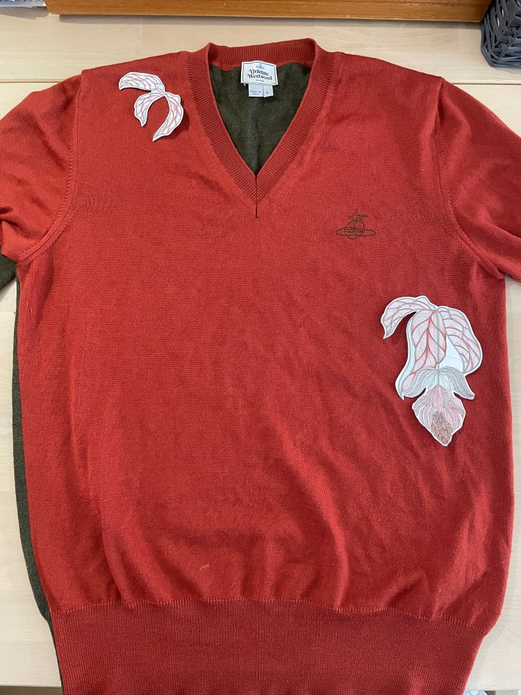 An orange Vivienne Westwood jumper lies on a table with paper embroidery designs placed onto it of a medinilla flower and its leaves to plan the placement of the stitching