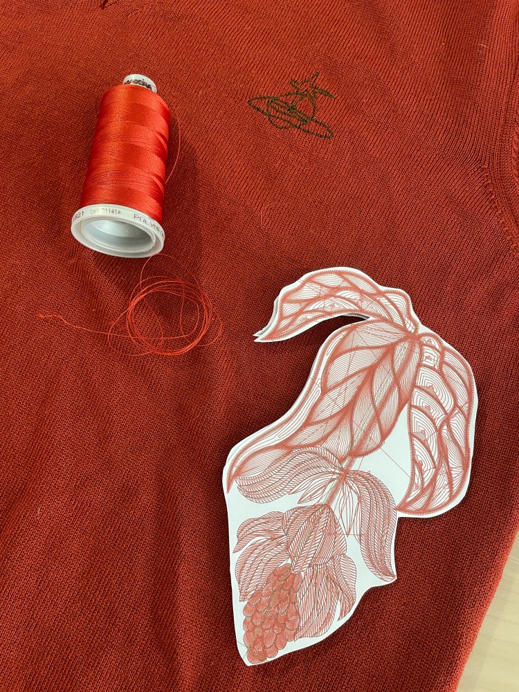 An orange spool of machine embroidery thread and a paper version of a Medinilla flower embroidery design lie on a fine knitted Vivienne Westwood jumper where the VW logo is visible in green