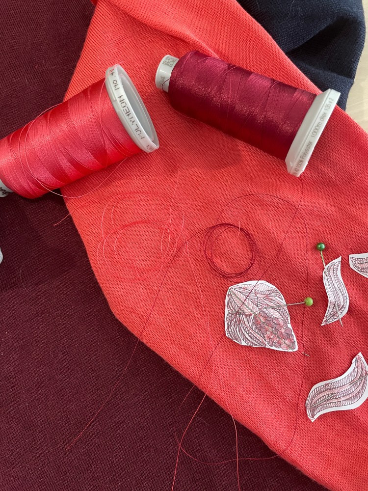Two machine embroidery threads lie on a pink knitted sleeve of a jumper with moth holes in it. Paper cut outs of the embroidery design are pinned into place over the moth holes
