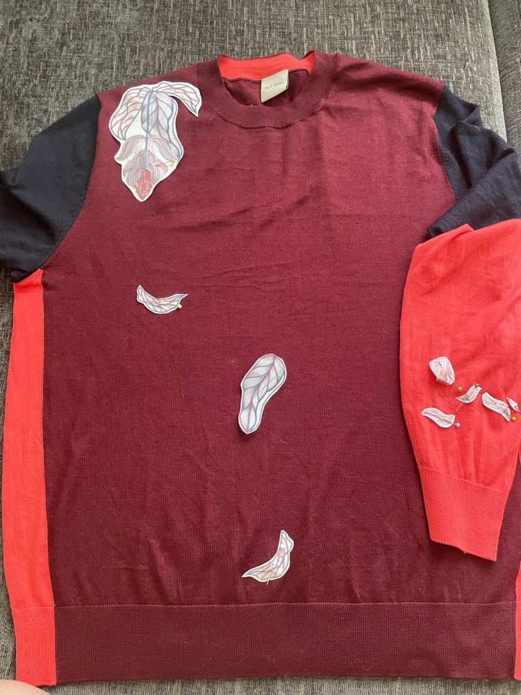 A Burgundy jumper with navy and pink sleeves lies on a grey background with paper medinilla flower designs pinned to it