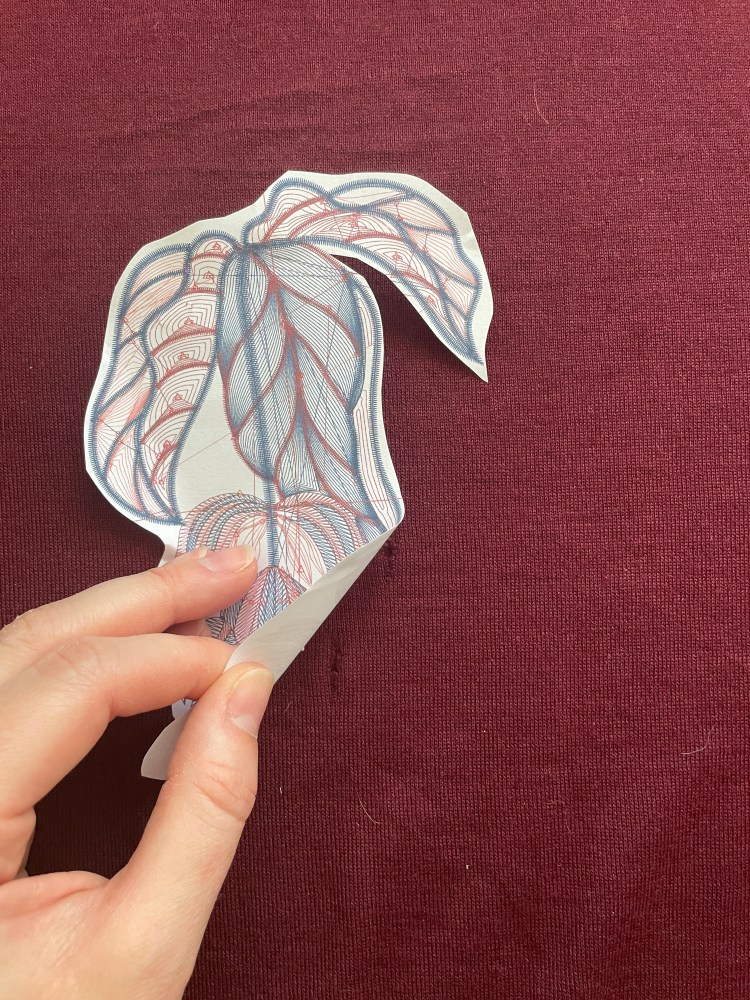 A hand peels back a cut out of an embroidery design placed on a burgundy knit fabric with a moth hole visible under the cut out
