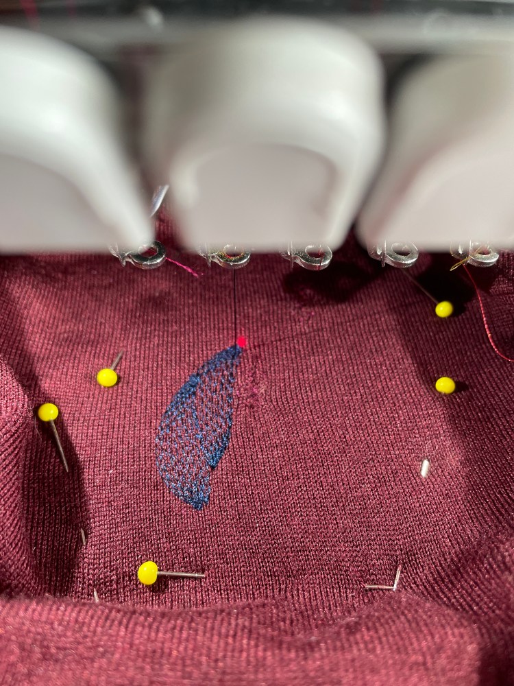 Burgundy knitted fabric pinned with yellow headed pins is being embroidered in an embroidery machine with navy blue thread in a leaf shape.