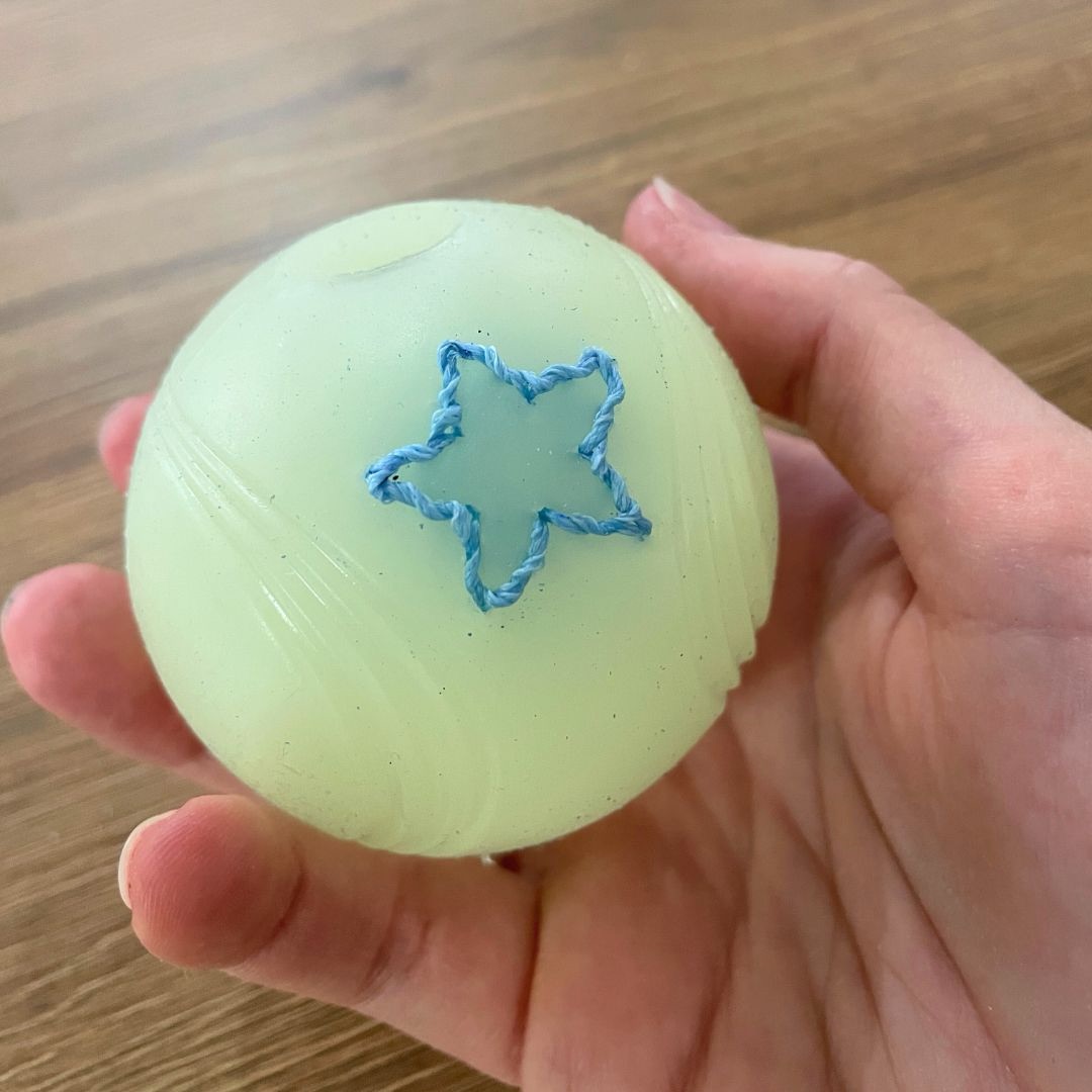 Pale green to white rubber dog ball with a blue embroidered star on it in and hand against a wooden background