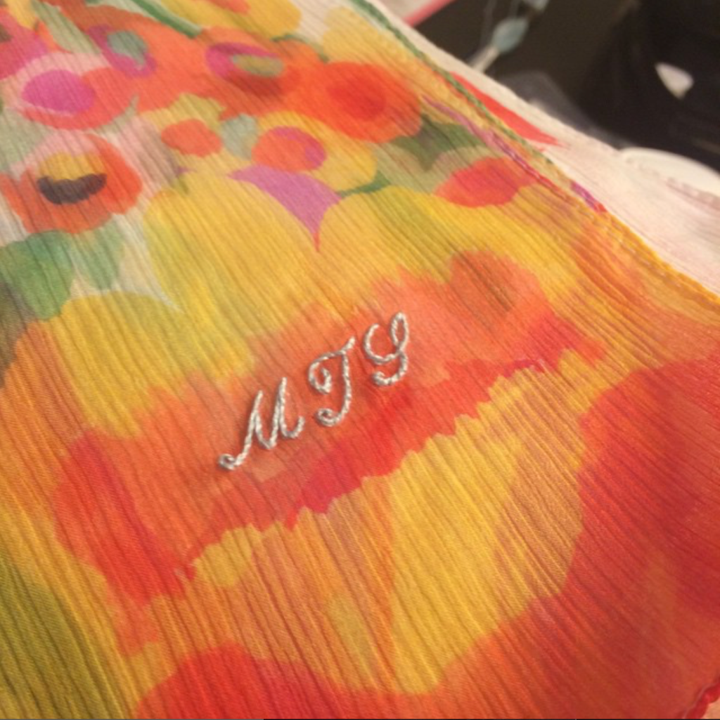 Light blue script monogram of MTS on a sheer orange and yellow patterned silk scarf